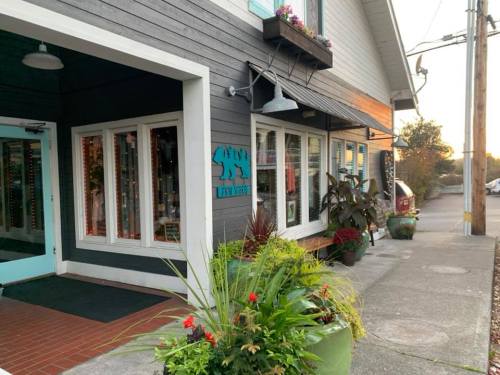 A storefront with a blue bear sign, surrounded by plants and flowers, on a quiet street during sunset.