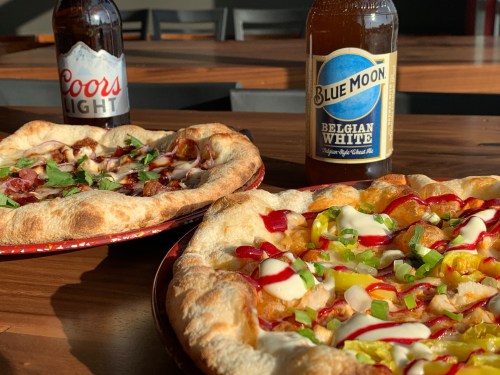 Two pizzas with various toppings and sauces, alongside bottles of Coors Light and Blue Moon beer on a wooden table.