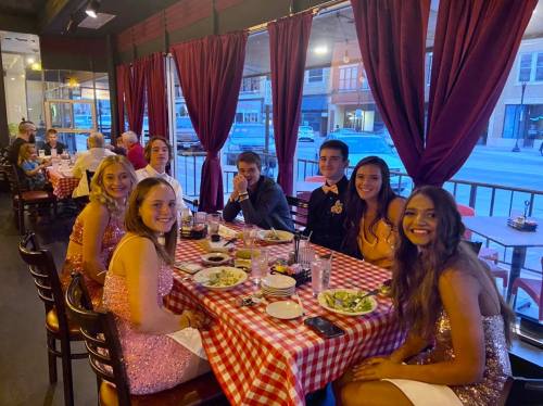 A group of seven friends in formal attire enjoying dinner at a restaurant with red checkered tablecloths.