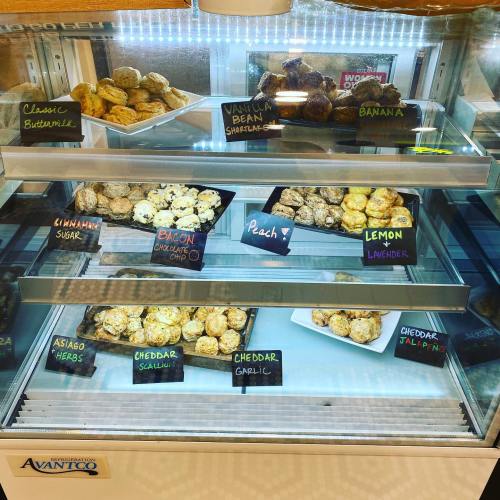 A display case filled with various baked goods, including muffins and scones, labeled with flavors like lemon and peach.