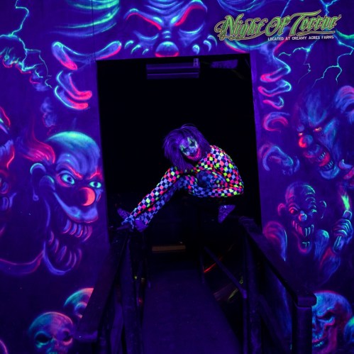 A performer in a colorful outfit poses in a dark, neon-lit hallway with eerie, glowing murals on the walls.