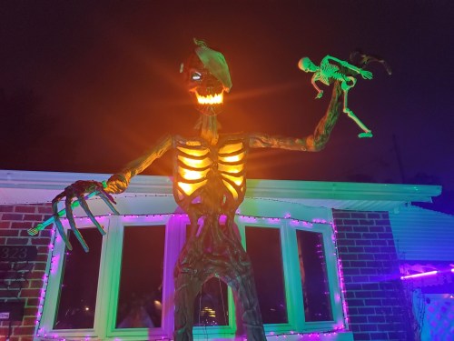 A large, illuminated skeleton figure holds a smaller skeleton, set against a dark night with colorful lights.