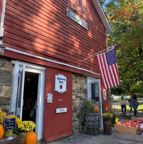 A red barn-style cider mill with an American flag, pumpkins, and flowers outside, welcoming visitors to the farm.