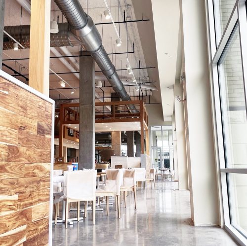 Modern restaurant interior with wooden accents, white chairs, and large windows, featuring an open and airy atmosphere.