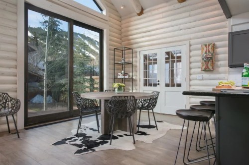 A cozy dining area with a round table, stylish chairs, and large windows overlooking a forested view.