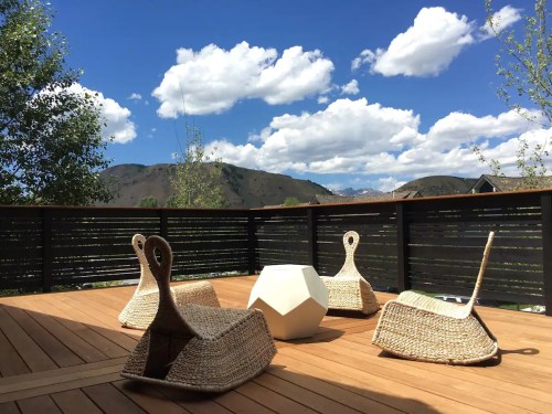 A sunny outdoor deck with woven chairs and a geometric table, surrounded by mountains and a blue sky with clouds.