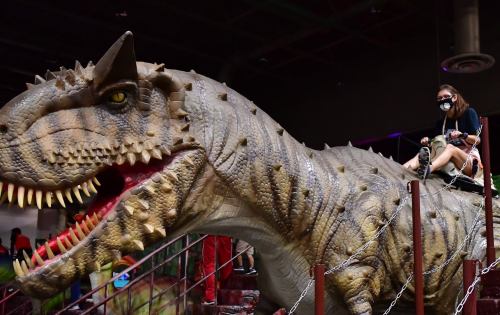 A person wearing a mask rides a large dinosaur statue in a colorful indoor setting.