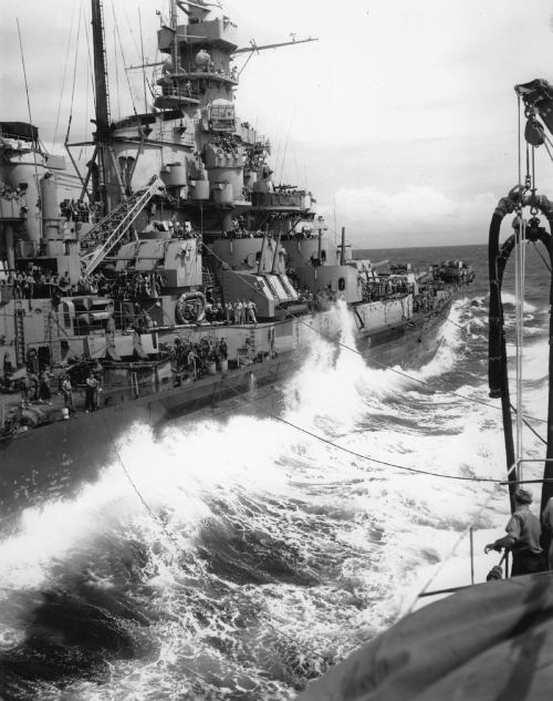 A battleship navigates through rough seas, with crew members visible on deck amidst crashing waves.