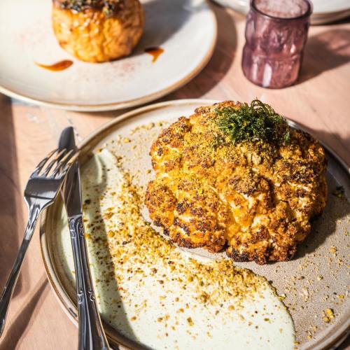A roasted cauliflower head topped with herbs and spices, served on a plate with a creamy sauce and crushed nuts.