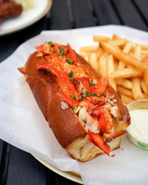 A lobster roll filled with lobster meat, garnished with herbs, served with a side of fries on a paper-lined plate.