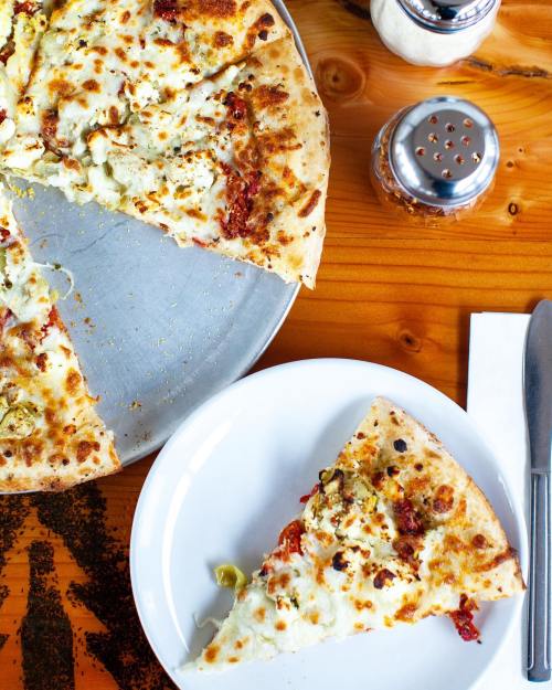 A pizza with toppings on a metal tray, with one slice on a white plate, set on a wooden table.