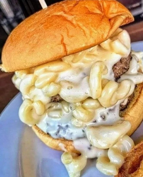 A close-up of a burger topped with creamy macaroni and cheese, overflowing from a toasted bun.
