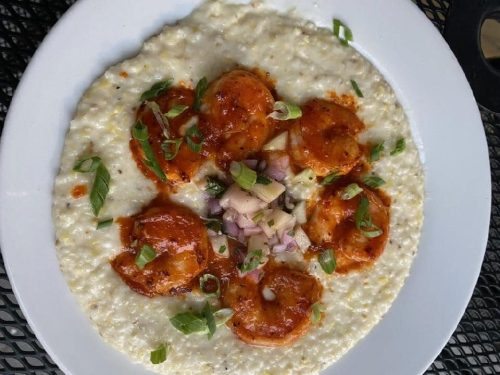A plate of creamy grits topped with six shrimp, garnished with green onions and a mix of diced vegetables.