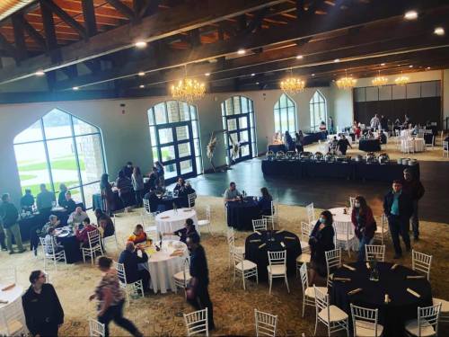 A spacious event hall with tables, chairs, and guests mingling, featuring large windows and chandeliers.