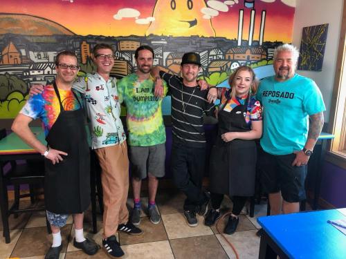 A group of six people in colorful shirts and aprons pose together in front of a vibrant mural.