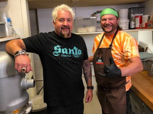 Two men smiling in a kitchen, one wearing a black shirt and the other in an orange tie-dye shirt and apron.