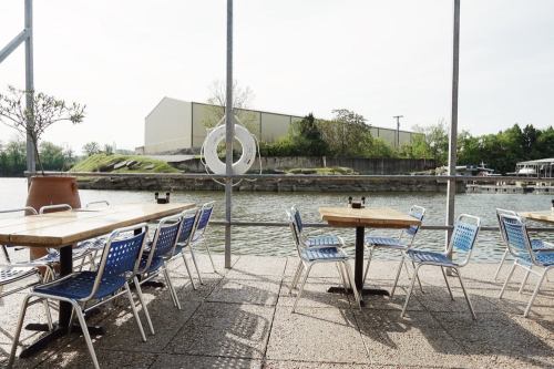 Outdoor seating area by a calm waterway, featuring wooden tables and blue chairs, with a building in the background.