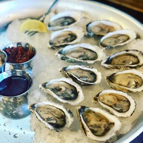 A platter of fresh oysters on ice, accompanied by two dipping sauces and a lemon wedge.