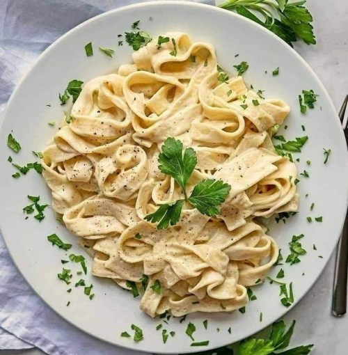 A plate of fettuccine pasta garnished with parsley and black pepper on a light background.