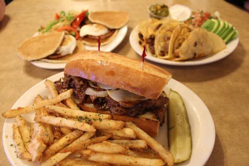 A hearty sandwich with fries and a pickle, alongside a plate of tacos and pancakes with salad in the background.