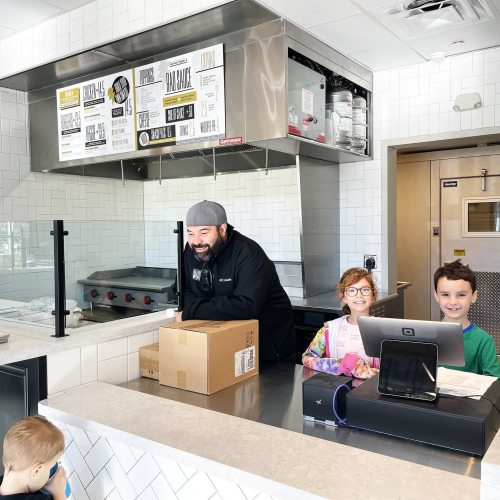 A smiling chef stands behind a counter with two children, one with glasses, at a casual eatery.