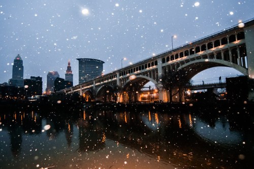 A snowy evening view of a bridge over a river, with city buildings illuminated in the background.