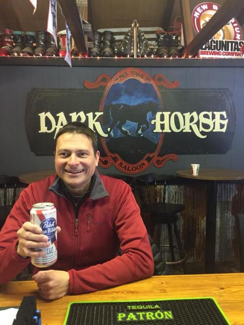 A smiling man in a red jacket holds a beer can at a bar with a "Dark Horse Saloon" sign in the background.