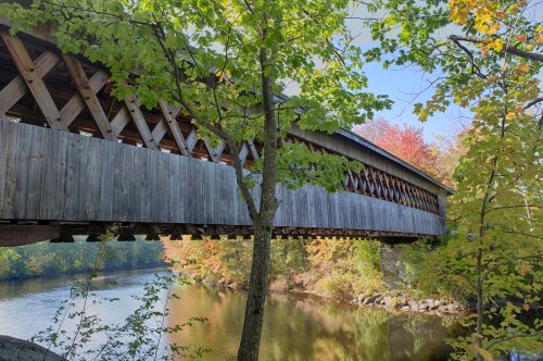 A wooden covered bridge spans a calm river, surrounded by vibrant autumn foliage and clear blue skies.