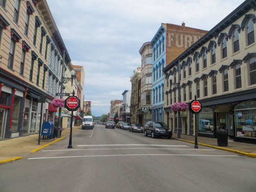 A quiet street lined with historic buildings, colorful storefronts, and flower baskets, with parked cars and a cloudy sky.