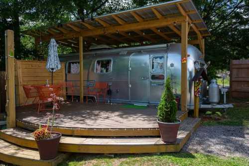 A silver Airstream trailer under a wooden pergola, surrounded by plants and outdoor seating on a deck.