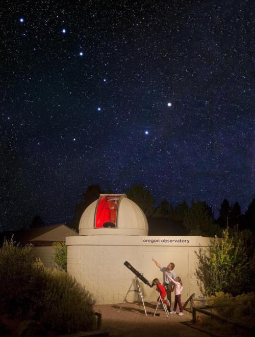 A person points at the night sky near a telescope outside the Oregon Observatory, with stars shining brightly above.