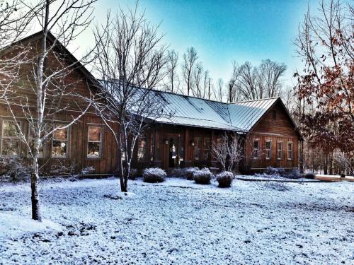 A wooden house surrounded by snow-covered ground and bare trees under a clear blue sky.