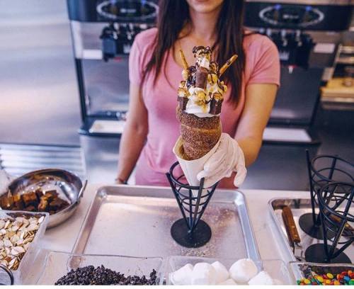 A woman holds a decorated ice cream cone topped with various sweets, standing behind a counter with toppings.
