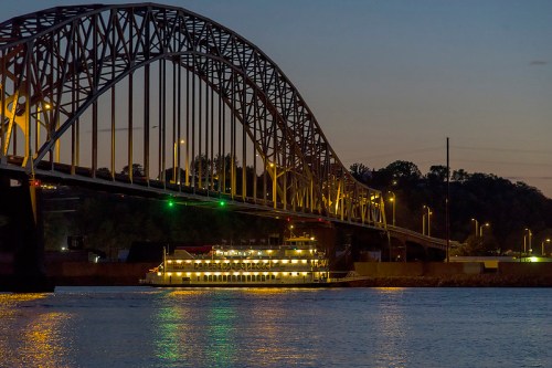 A lit boat passes under a large bridge at dusk, reflecting on the calm water below.