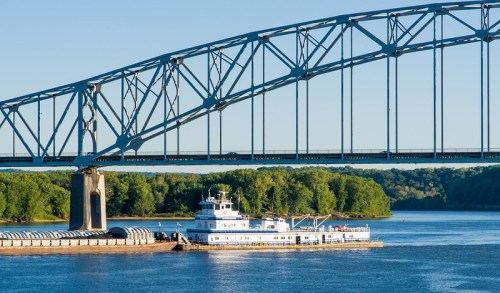 A barge docked under a large steel bridge, with calm water and green trees in the background.