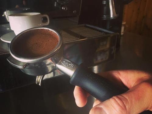 A hand holding a portafilter filled with coffee grounds, with a coffee machine and cup in the background.