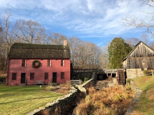A pink historic house with a wreath, next to a wooden bridge and a mill, surrounded by trees and a grassy area.