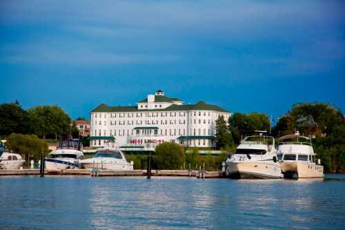 A waterfront view of a white hotel with green accents, surrounded by boats docked in the calm water.