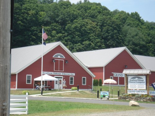 Red barn-style building with a flag, sign for Grafton Village Cheese Co., and green hills in the background.
