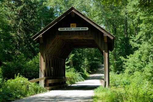 A wooden covered bridge surrounded by lush greenery, leading to a dirt path. Sign reads "Swamp Meadow Covered Bridge."