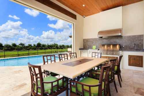 Outdoor dining area with a large table and chairs, overlooking a pool and green landscape under a blue sky.