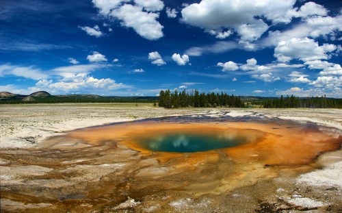 A vibrant hot spring surrounded by colorful mineral deposits under a blue sky with fluffy clouds.