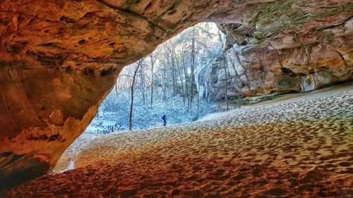 A person stands in a sandy cave entrance, framed by rocky walls and a snowy forest in the background.