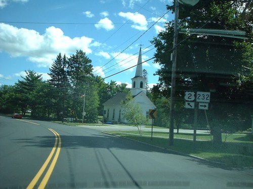 A winding road leads to a white church with a steeple, surrounded by trees and blue skies. Road signs indicate directions.