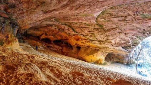 A person stands inside a large, colorful cave with sandy ground and textured rock walls.