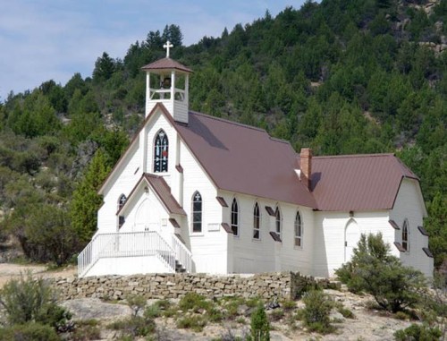 A quaint white church with a bell tower, surrounded by green trees and hills under a cloudy sky.
