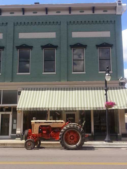 A vintage red tractor parked in front of a green brick building with large windows and a striped awning.