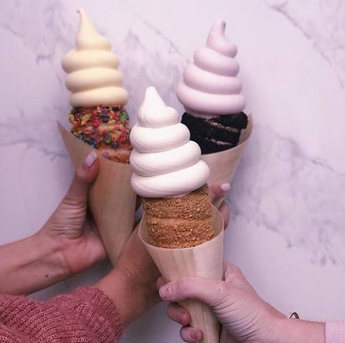 Three hands holding ice cream cones with swirled soft serve in various flavors against a marble background.