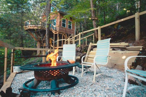 A cozy fire pit with chairs in front of a treehouse, surrounded by trees and gravel pathways.