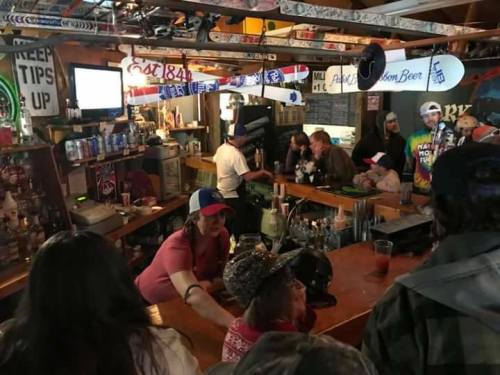 A lively bar scene with patrons socializing, a bartender serving drinks, and ski equipment decor overhead.